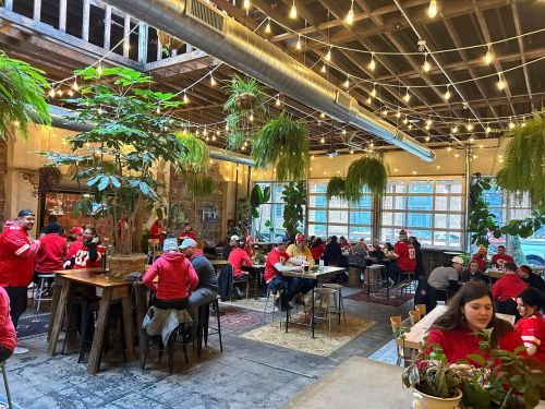 A lively restaurant filled with patrons in red attire, surrounded by plants and warm string lights.