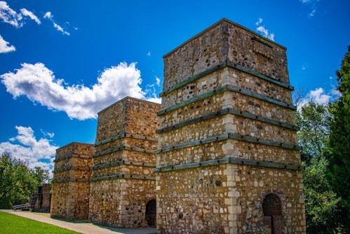 Three stone towers with a blue sky and fluffy clouds in the background, surrounded by green grass and trees.
