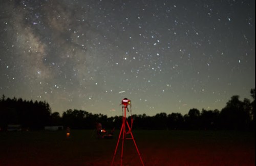 A tripod with a red light stands in a field under a starry night sky, featuring the Milky Way.