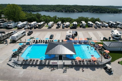 Aerial view of a pool area with lounge chairs and umbrellas, surrounded by RVs and a lake in the background.