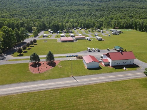 Aerial view of a campground with RVs, grassy areas, and a few buildings along a road surrounded by trees.