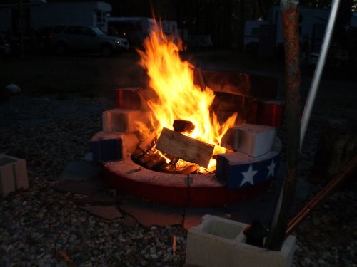 A vibrant campfire burns in a stone fire pit, surrounded by gravel and trees in the background.