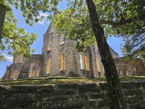 Ruins of a stone building surrounded by trees, with blue sky and sunlight filtering through the leaves.