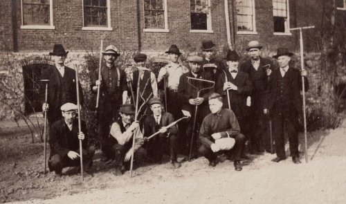 A group of men in historical clothing, holding tools, pose together outdoors near a brick building.