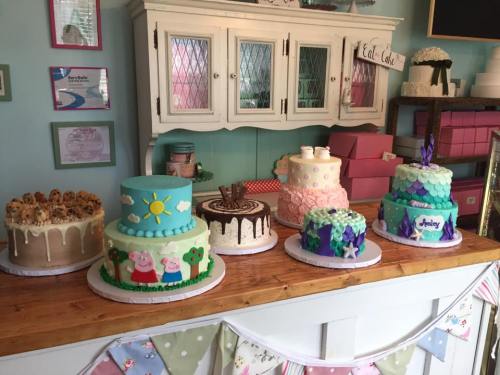 A display of colorful cakes on a wooden table, featuring various designs and decorations in a charming bakery setting.