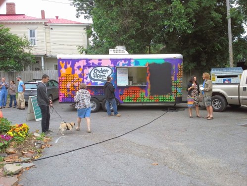 A colorful food truck with people ordering, a dog on a leash, and a vibrant outdoor setting with flowers.