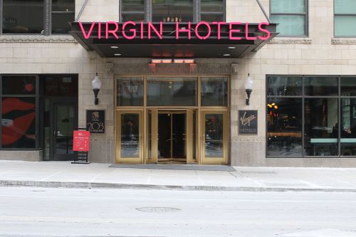 Entrance of Virgin Hotels with large glass doors and a neon sign, located in an urban setting.