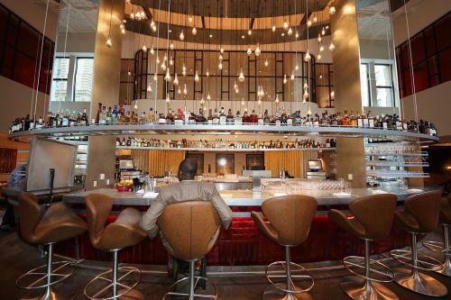 A modern bar with a curved counter, high stools, and shelves lined with various bottles, illuminated by hanging lights.