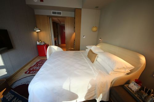 A modern hotel room featuring a large bed, wooden accents, and a view of an open doorway leading to another area.