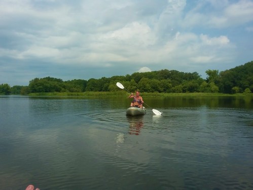 A person paddles a kayak on a calm lake surrounded by lush greenery under a partly cloudy sky.
