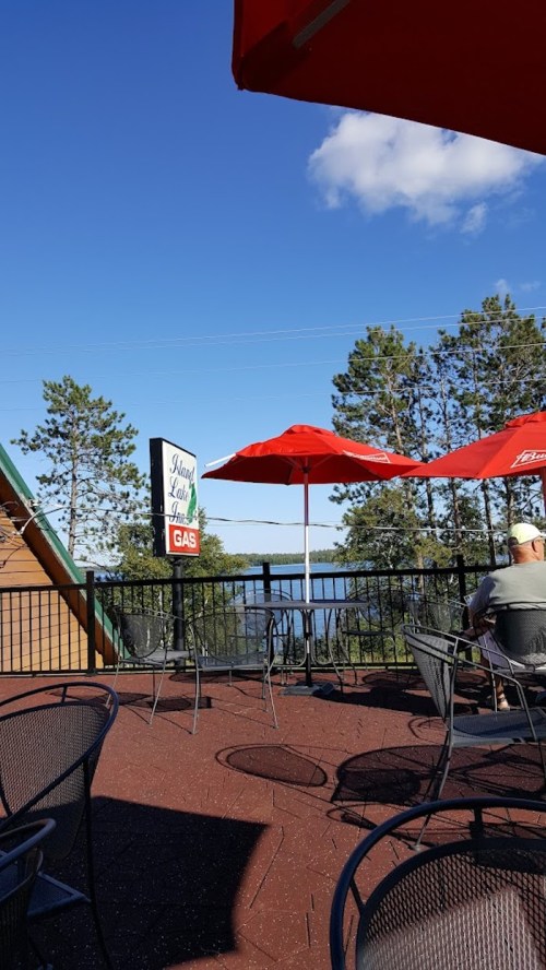 A scenic outdoor patio with red umbrellas, overlooking a lake and trees under a clear blue sky.