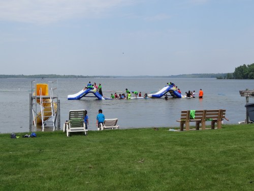 Children playing on inflatable slides in a lake, with benches and a lifeguard station nearby.