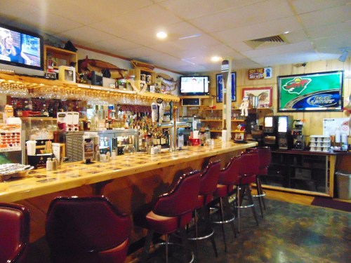 A cozy bar interior with a wooden counter, high stools, and multiple TVs displaying sports. Decorated with memorabilia.
