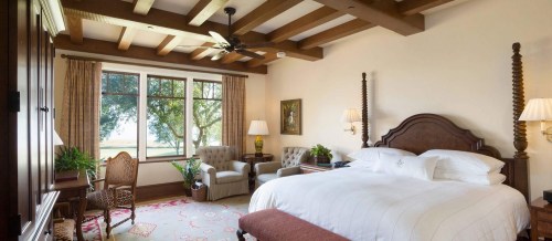 Cozy bedroom with a large bed, armchairs, wooden beams, and a view of trees through a large window.