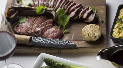 A wooden platter with sliced steak, roasted garlic, asparagus, and a glass of red wine.