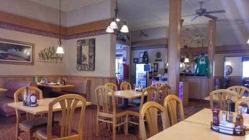 Cozy restaurant interior with wooden tables, chairs, and decor, featuring booths and a counter in the background.