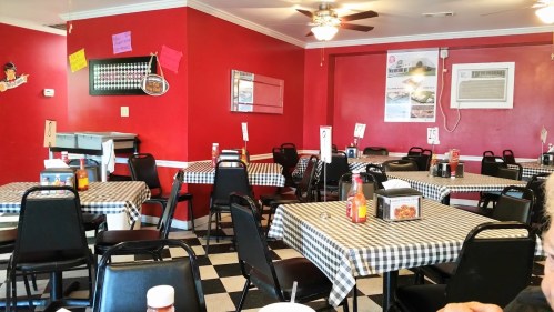 A cozy diner with red walls, checkered tablecloths, and empty black chairs, featuring condiments on the tables.