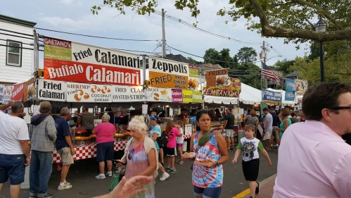 A bustling food festival with various stalls selling fried calamari, zeppolis, and other treats, surrounded by crowds.
