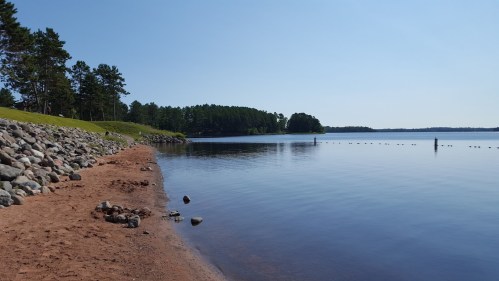 A serene lakeside view with a sandy shore, smooth water, and trees lining the horizon under a clear blue sky.