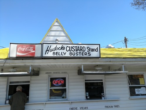 A small custard stand with a yellow roof, featuring a sign that reads "Hudock Custard Stand Belly Busters" and an open sign.
