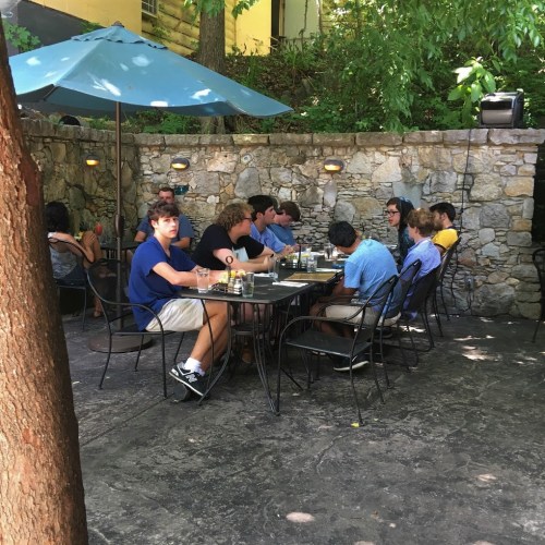 A group of people dining outdoors under an umbrella, surrounded by trees and a stone wall.