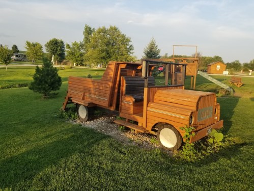 A wooden truck sculpture in a grassy park, surrounded by trees and a clear blue sky.