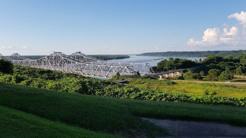 A scenic view of a bridge spanning a river, surrounded by lush greenery and a clear blue sky.