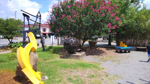 A park scene featuring colorful sculptures, greenery, and a blue bench under a bright sky.
