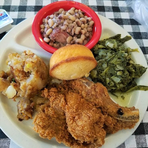 A plate of fried chicken, cornbread, collard greens, black-eyed peas, and seasoned potatoes on a checkered tablecloth.