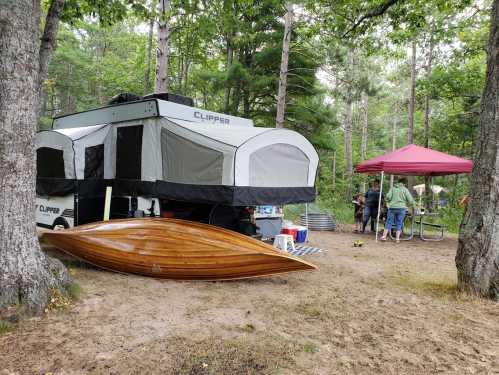 A pop-up camper in a wooded area, with a wooden boat nearby and people gathered under a red canopy.