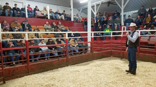 A speaker in a cowboy hat addresses a seated audience in a livestock auction arena, surrounded by wooden railings.