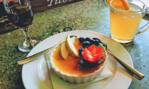 A creamy dessert topped with fruit, served with a glass of juice and a wine glass on a green table.
