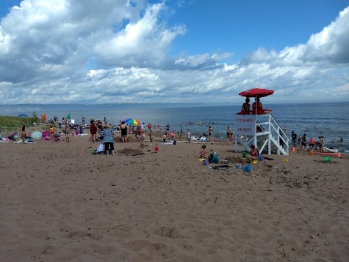 A sandy beach scene with people sunbathing, swimming, and a lifeguard tower under a blue sky with clouds.