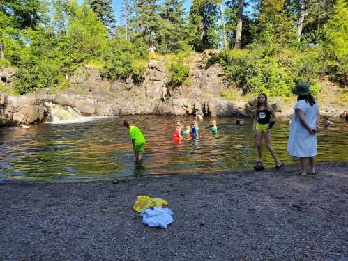 A group of children play in a natural pool while adults watch from the shore, surrounded by trees and rocks.