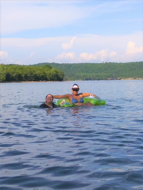 Two women enjoy a sunny day on a lake, one floating on a green inflatable while the other swims nearby.