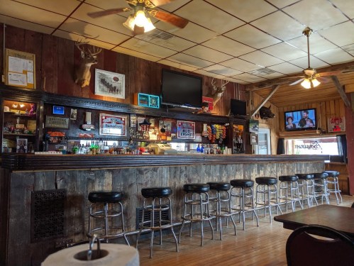A rustic bar with wooden decor, stools, and a TV, featuring beer signs and mounted deer heads on the walls.