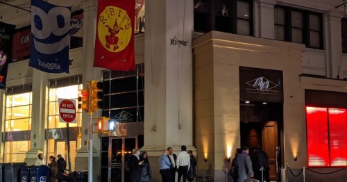 A city street scene at night featuring a building entrance with flags and people gathered outside.