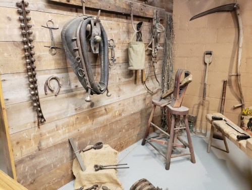 A rustic display of vintage farming tools and equipment hanging on a wooden wall, with a stool and workbench nearby.