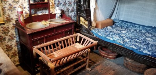 A vintage room featuring a wooden sink, a cradle, and a bed with blue bedding against floral wallpaper.