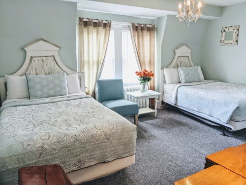 Cozy hotel room with two beds, a blue chair, and a small table, featuring soft lighting and neutral decor.