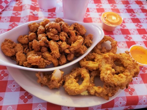 A plate of fried seafood, including shrimp and onion rings, served with dipping sauces on a checkered tablecloth.