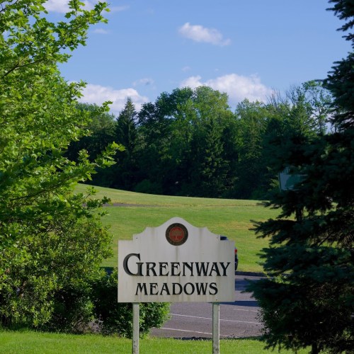 Sign for Greenway Meadows with a grassy field and trees in the background on a sunny day.