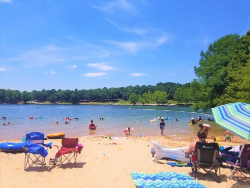 A sunny beach scene with people swimming, lounging on chairs, and enjoying the water under a clear blue sky.