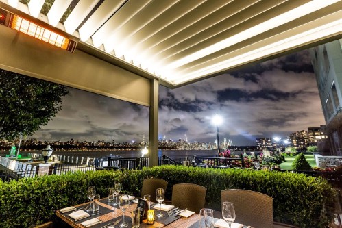 A beautifully lit outdoor dining area with a view of a city skyline at night, surrounded by greenery and soft lighting.