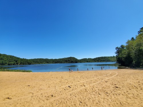 A sandy beach by a calm lake, surrounded by green hills under a clear blue sky, with people enjoying the water.