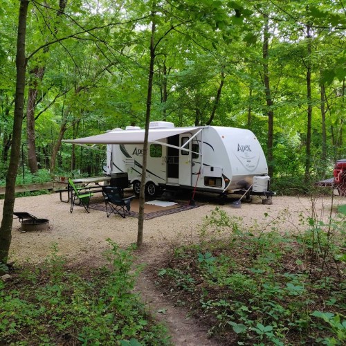 A camper parked in a wooded area, surrounded by trees and a gravel path, with outdoor seating nearby.