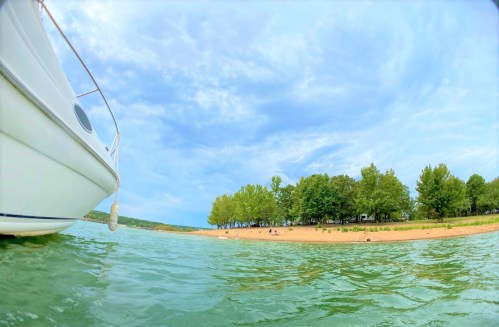 A boat floats in clear water near a sandy beach lined with green trees under a blue sky with scattered clouds.