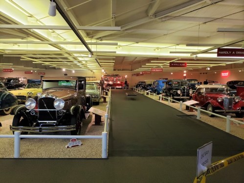 A vintage car museum featuring a variety of classic cars displayed on a carpeted floor under bright lights.