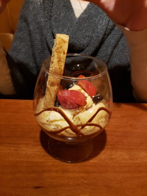 A dessert glass filled with ice cream, topped with strawberries, blueberries, chocolate drizzle, and a cookie stick.