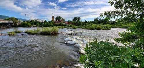 A scenic river with gentle rapids, surrounded by greenery and buildings under a blue sky with scattered clouds.